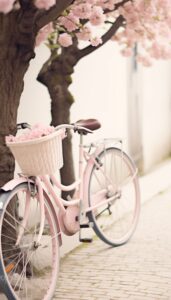 A pastel pink bicycle with a wicker basket filled with cherry blossoms under a blooming tree, set against a cobblestone path for an iPhone wallpaper.