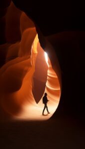 A person in silhouette stands inside a grand sandstone canyon, with light from above highlighting the smooth, flowing rock walls.